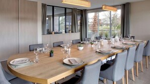 Uma sala de jantar moderna com uma mesa de madeira comprida posta para uma refeição, janelas grandes e vista para o exterior.