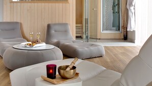 Um spa moderno com duas poltronas cinzentas, uma mesinha redonda com uma travessa de frutas e copos, uma tigela de madeira com um pilão, e uma área de chuveiro em vidro.