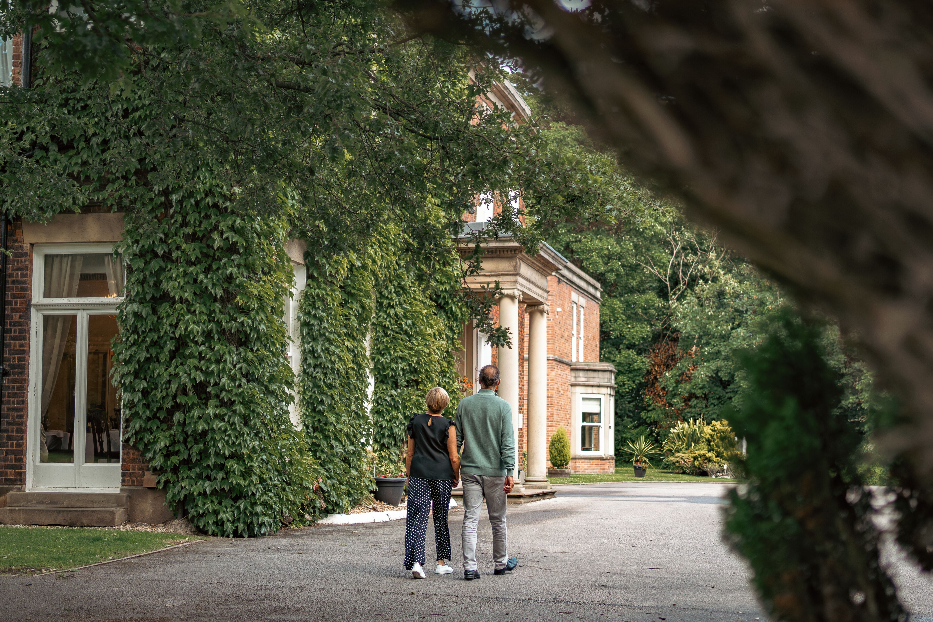 Un couple se tient devant un bâtiment à la façade en briques et aux colonnes, entouré de verdure.