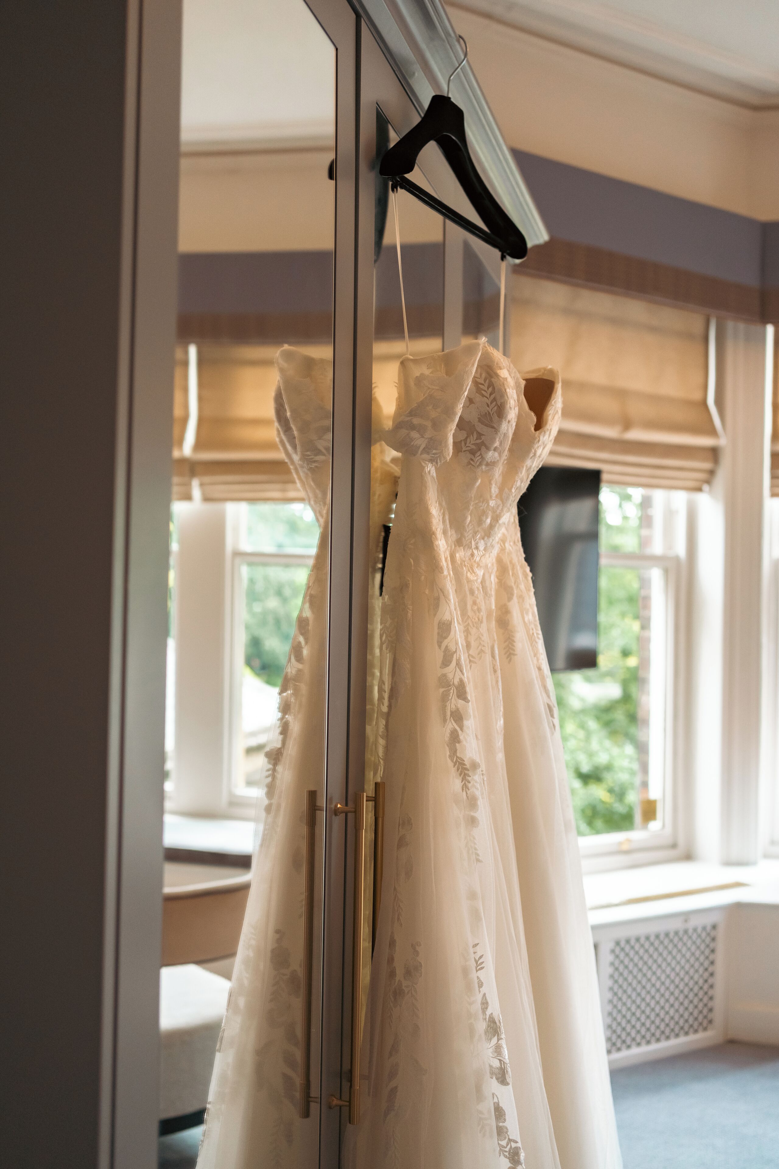 Un vestido de novia colgado de una percha en una habitación con ventanas amplias.