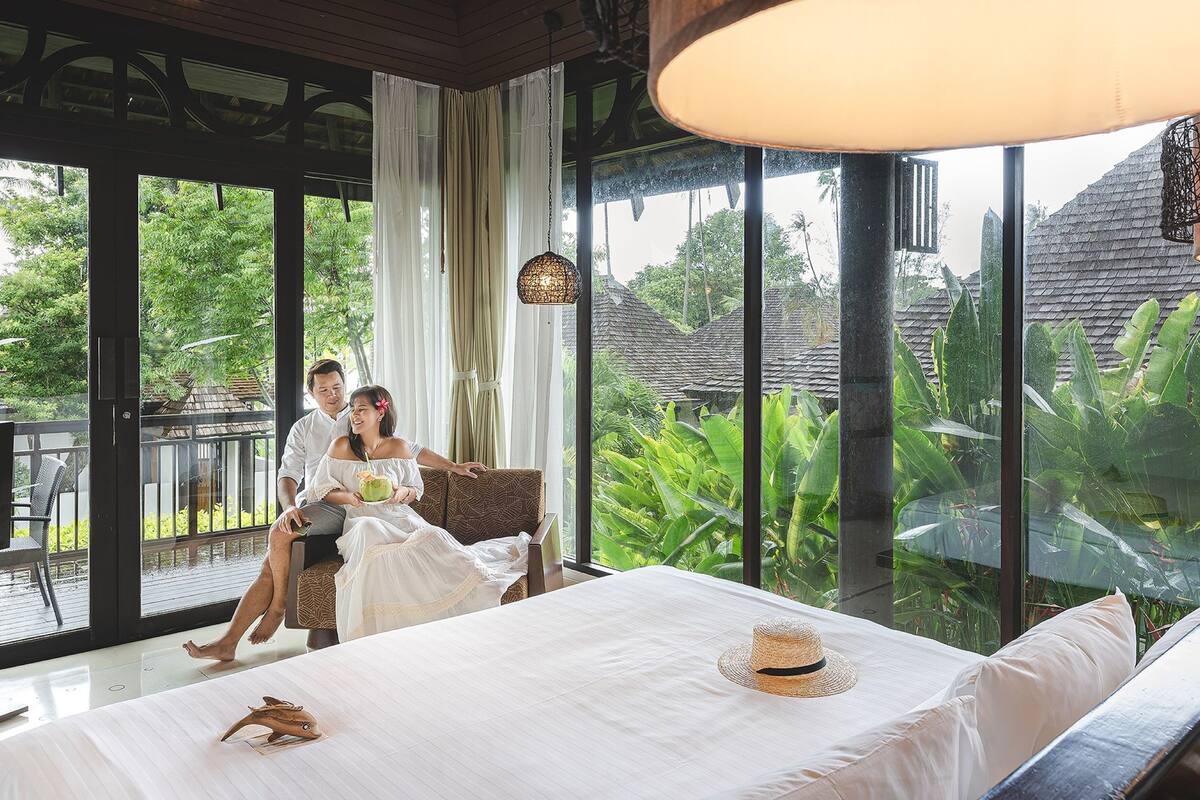 A bedroom with a large window, a bed with a straw hat, and a view of tropical vegetation.