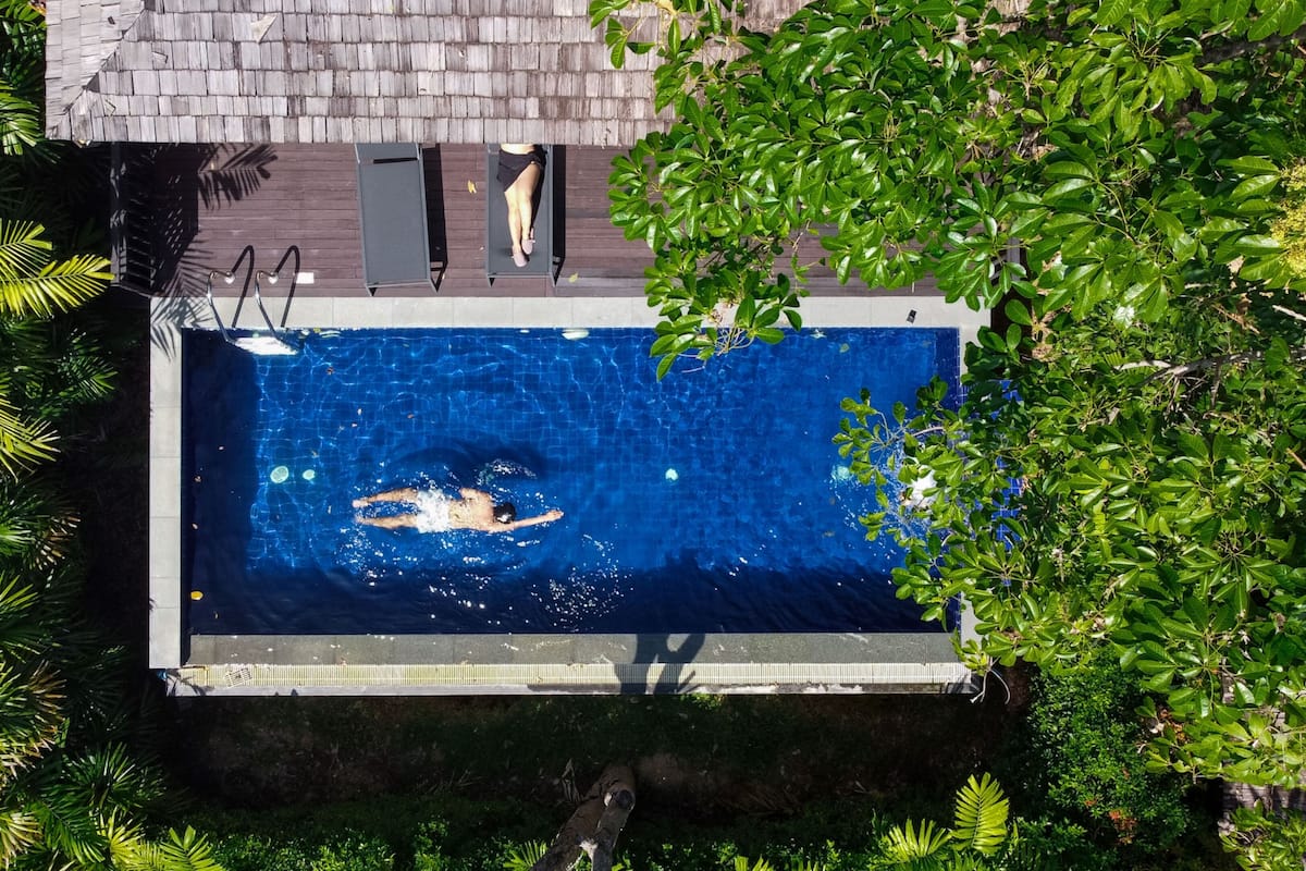 An aerial view of a rectangular swimming pool surrounded by lush greenery, with a wooden deck and a small window above the pool.
