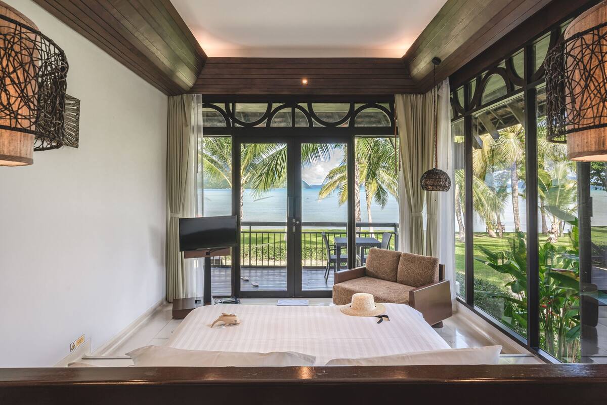 A bedroom with a large bed, a TV, and a view of a tropical landscape through large windows.