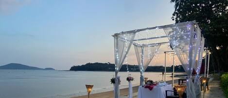 A beachside wedding setup with a canopy, tables, and a heart-shaped arrangement of rose petals and lights.