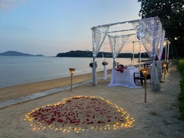 A beachside wedding setup with a canopy, tables, and a heart-shaped arrangement of rose petals and lights.