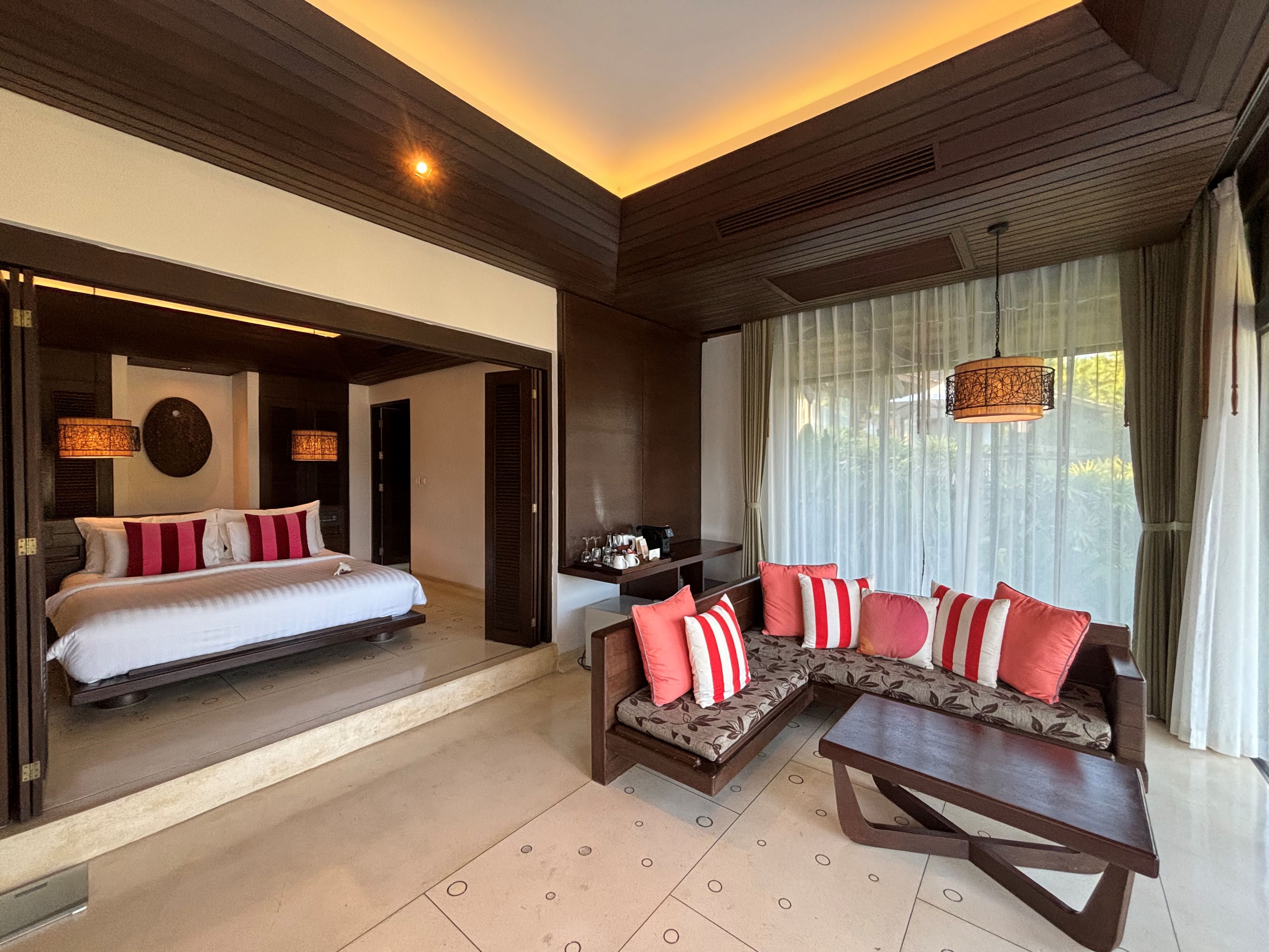 A modern hotel room with a large bed, a sofa, and a coffee table. There is a window with curtains and a view of greenery outside.