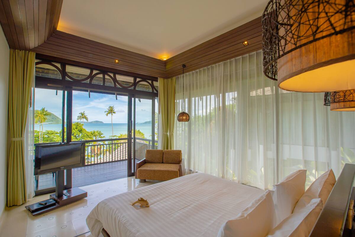 A bedroom with a large bed, a TV, a chair, and a balcony with a view of the sea.