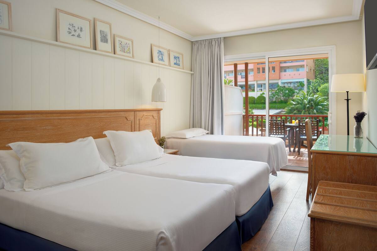 A hotel room with two beds, a wooden headboard, a balcony with a table and chairs, and framed pictures on the wall.
