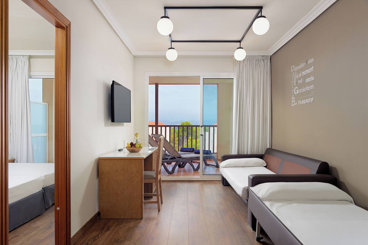 A hotel room with a bed, a desk, a chair, a TV, and a balcony with a view.