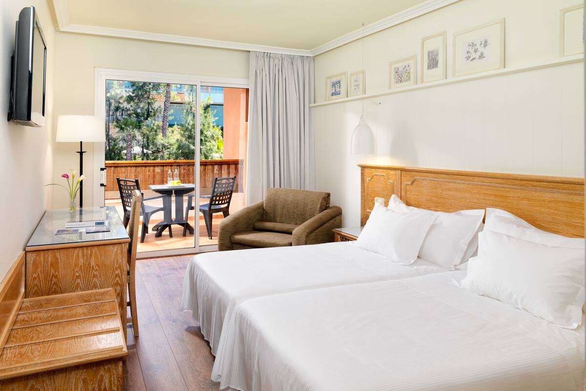 A hotel room with two beds, a desk with a lamp, a chair, and a balcony with a view of trees and buildings.