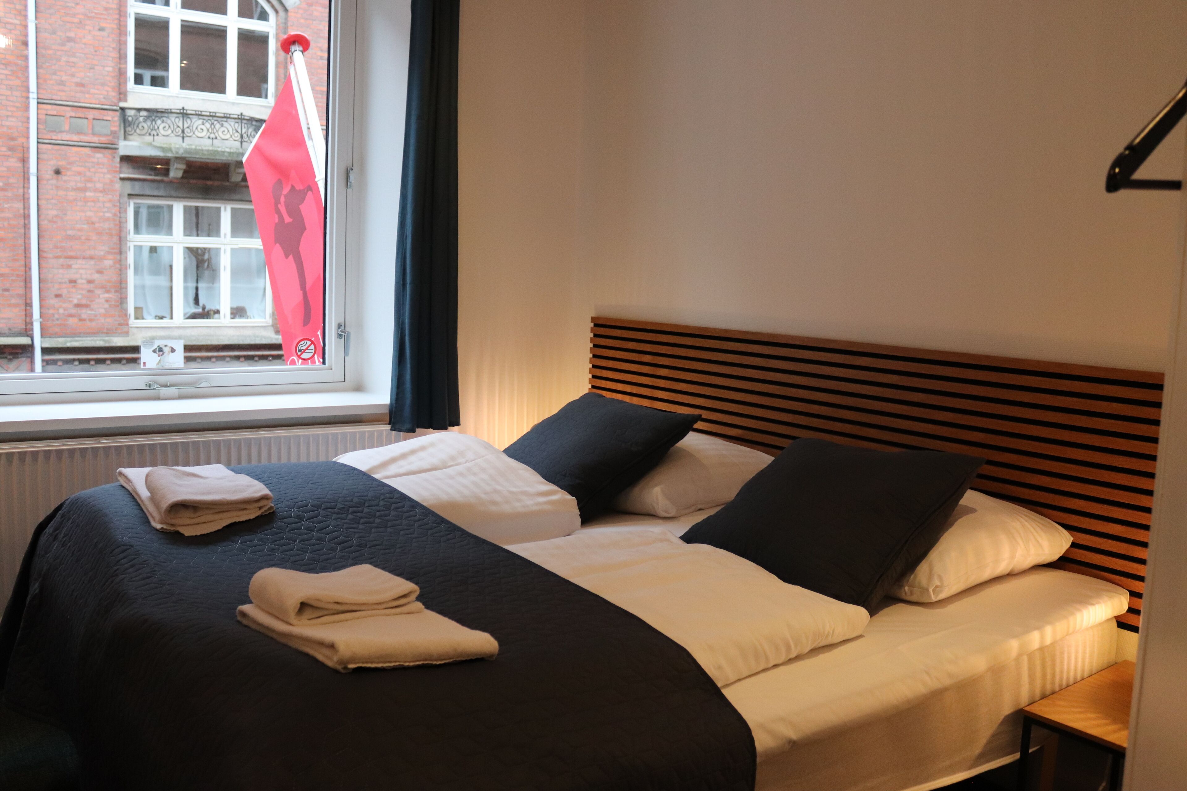Una cama bien hecha con edredón blanco y negro, cabecera de madera y ventana con vista a un edificio y una bandera roja.