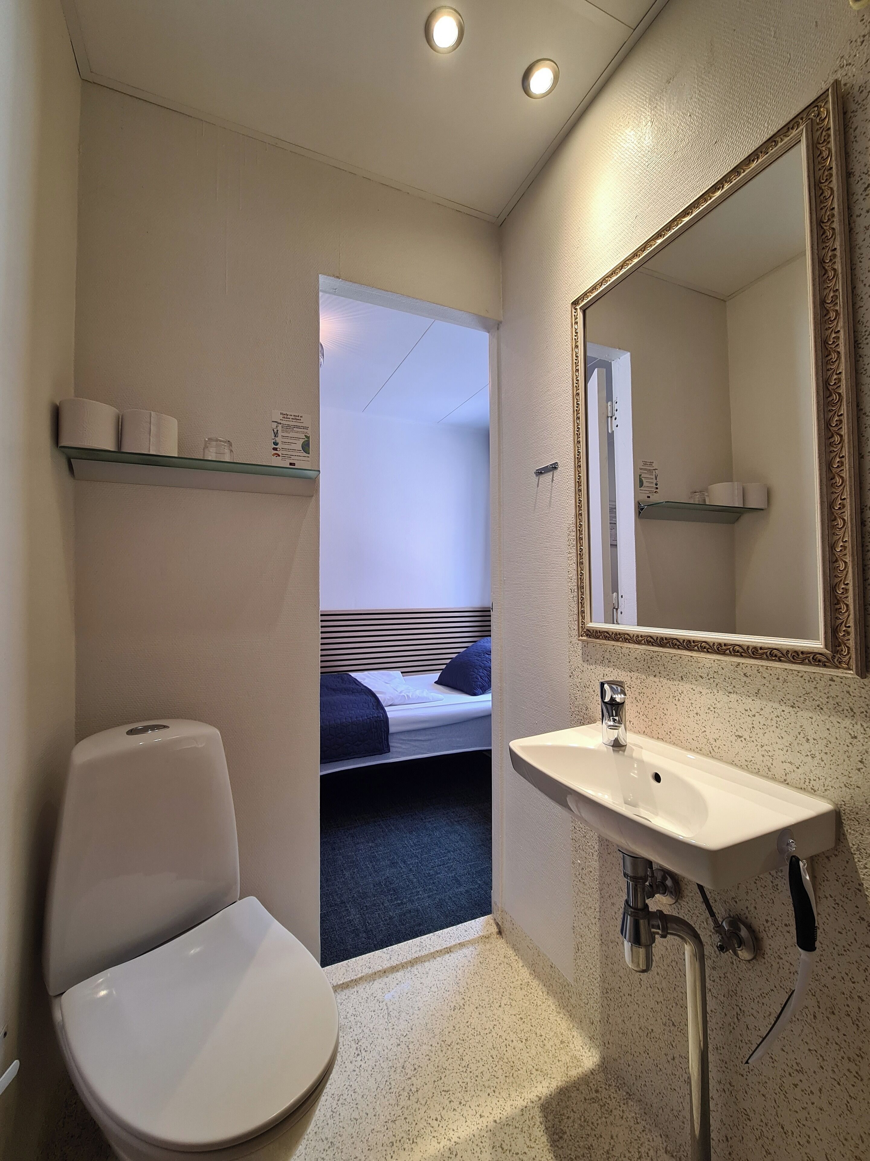 Baño pequeño y funcional con inodoro, lavabo y espejo. Hay un estante pequeño sobre el inodoro y uno más grande sobre el lavabo.