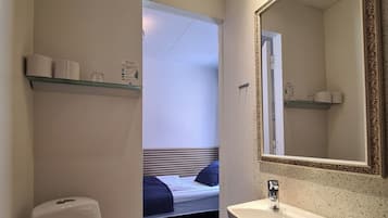 Baño pequeño y funcional con inodoro, lavabo y espejo. Hay un estante pequeño sobre el inodoro y uno más grande sobre el lavabo.