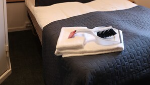 Un lit bien fait, recouvert d’une couverture sombre, avec deux oreillers noirs et un drap blanc. Deux lampes murales sont fixées de chaque côté du lit. Une serviette blanche et une brosse à cheveux sont posées sur le lit.