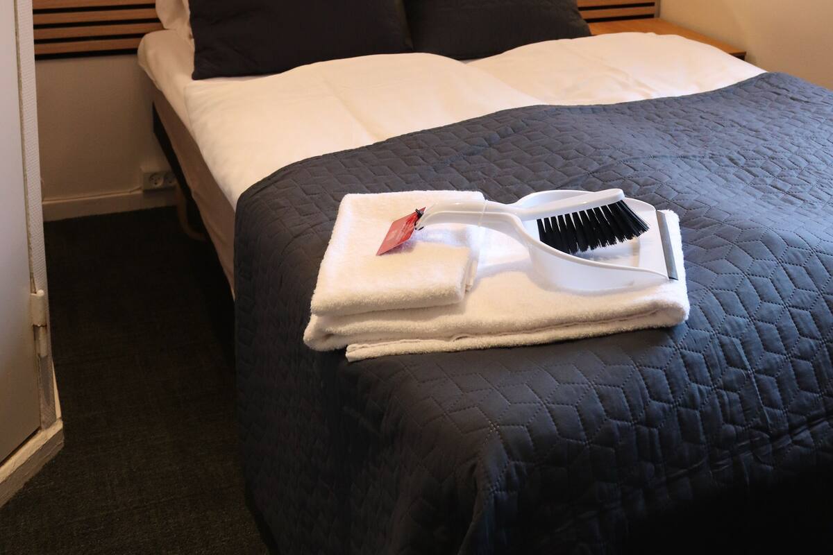 Una cama bien hecha con un edredón oscuro, dos almohadas negras y una manta blanca. Hay dos lámparas de pared a cada lado de la cama. Una toalla blanca y un cepillo para el cabello están sobre la cama.
