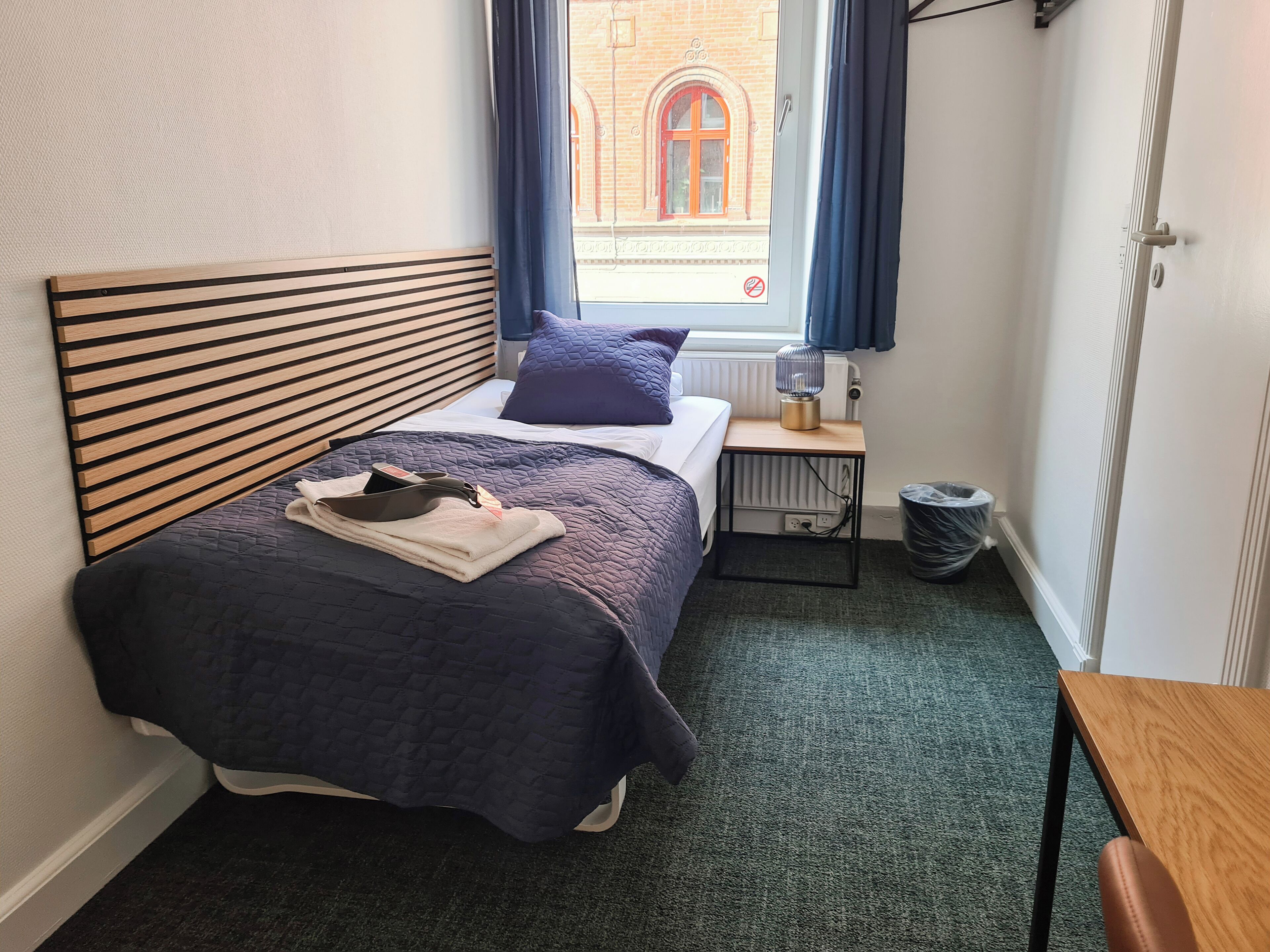 A neatly made bed with a headboard, a bedside table with a lamp, and a view of a building through a window.