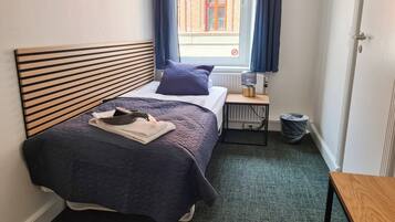 A neatly made bed with a headboard, a bedside table with a lamp, and a view of a building through a window.
