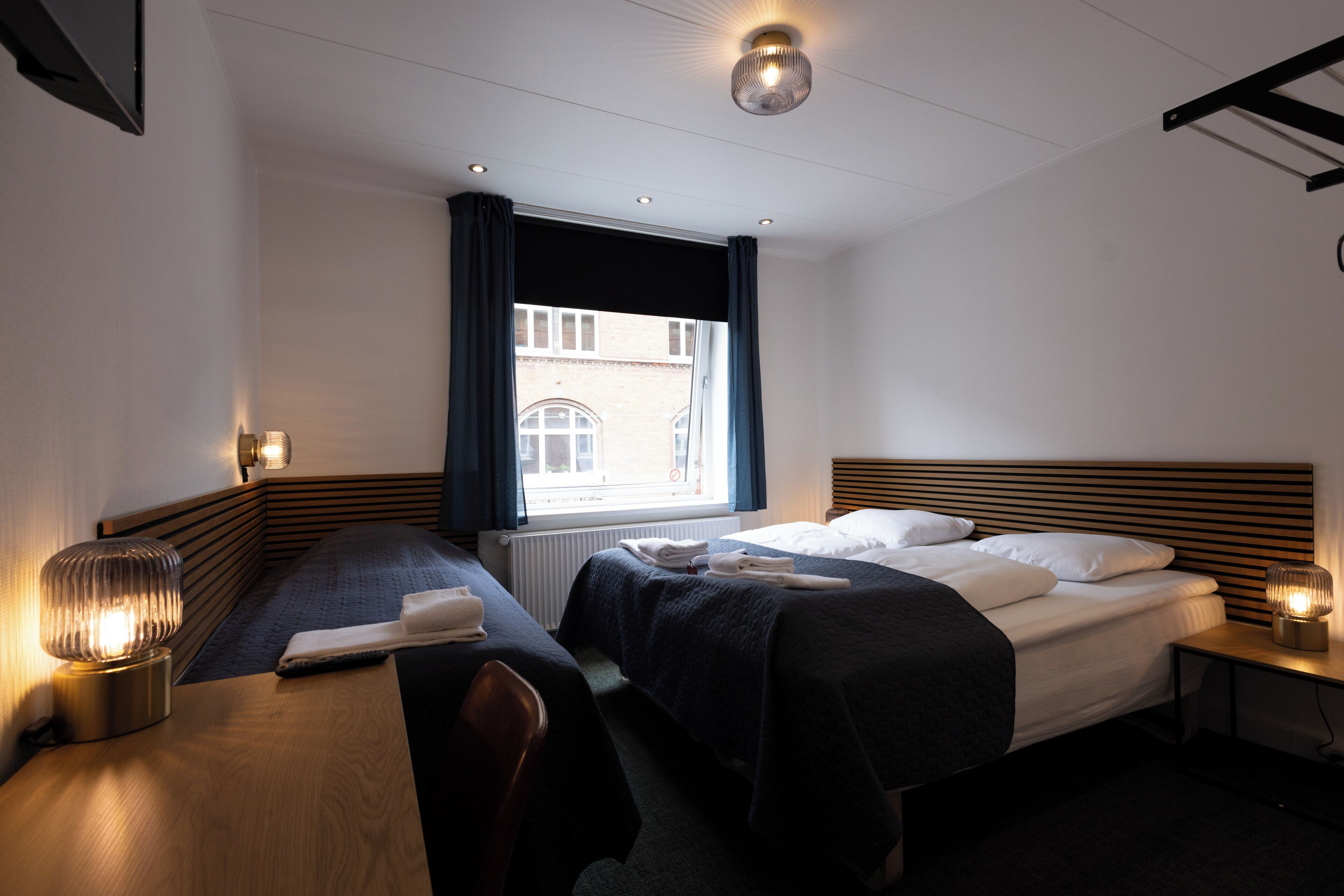 A hotel room with two beds, a wooden headboard, a window with curtains, a desk with a lamp, and a nightstand with a lamp.