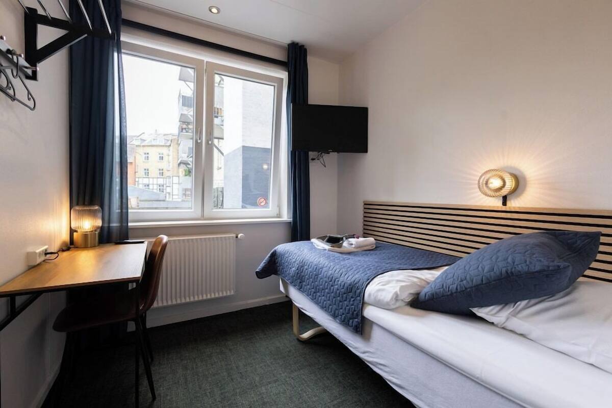 Habitación de hotel con cama, escritorio, silla, televisor y ventana con cortinas.