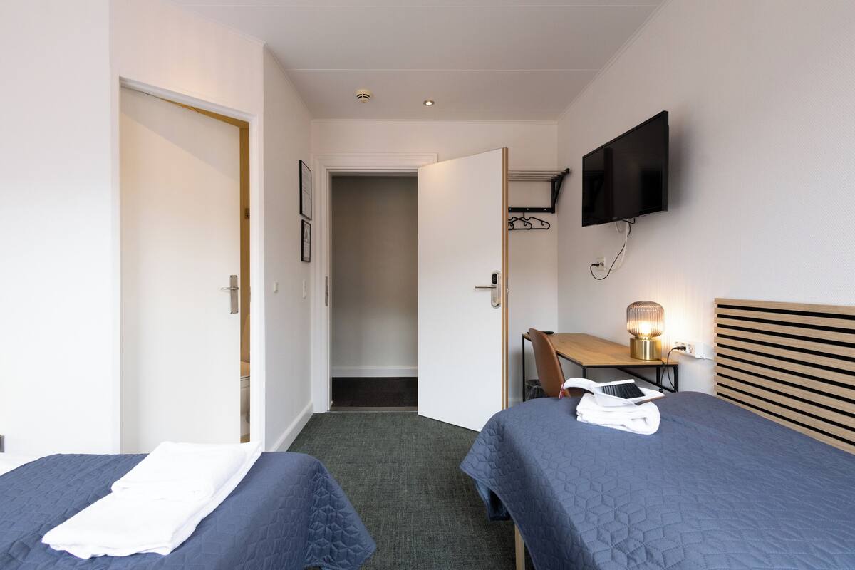 Habitación de hotel con dos camas, un escritorio con lámpara, televisión montada en la pared y una puerta que da al baño.