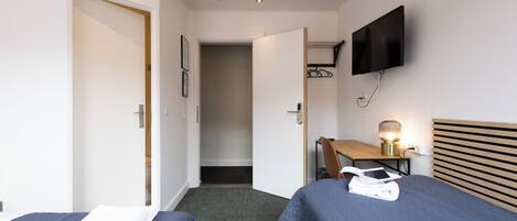 A hotel room with two beds, a desk with a lamp, a TV mounted on the wall, and a door leading to a bathroom.