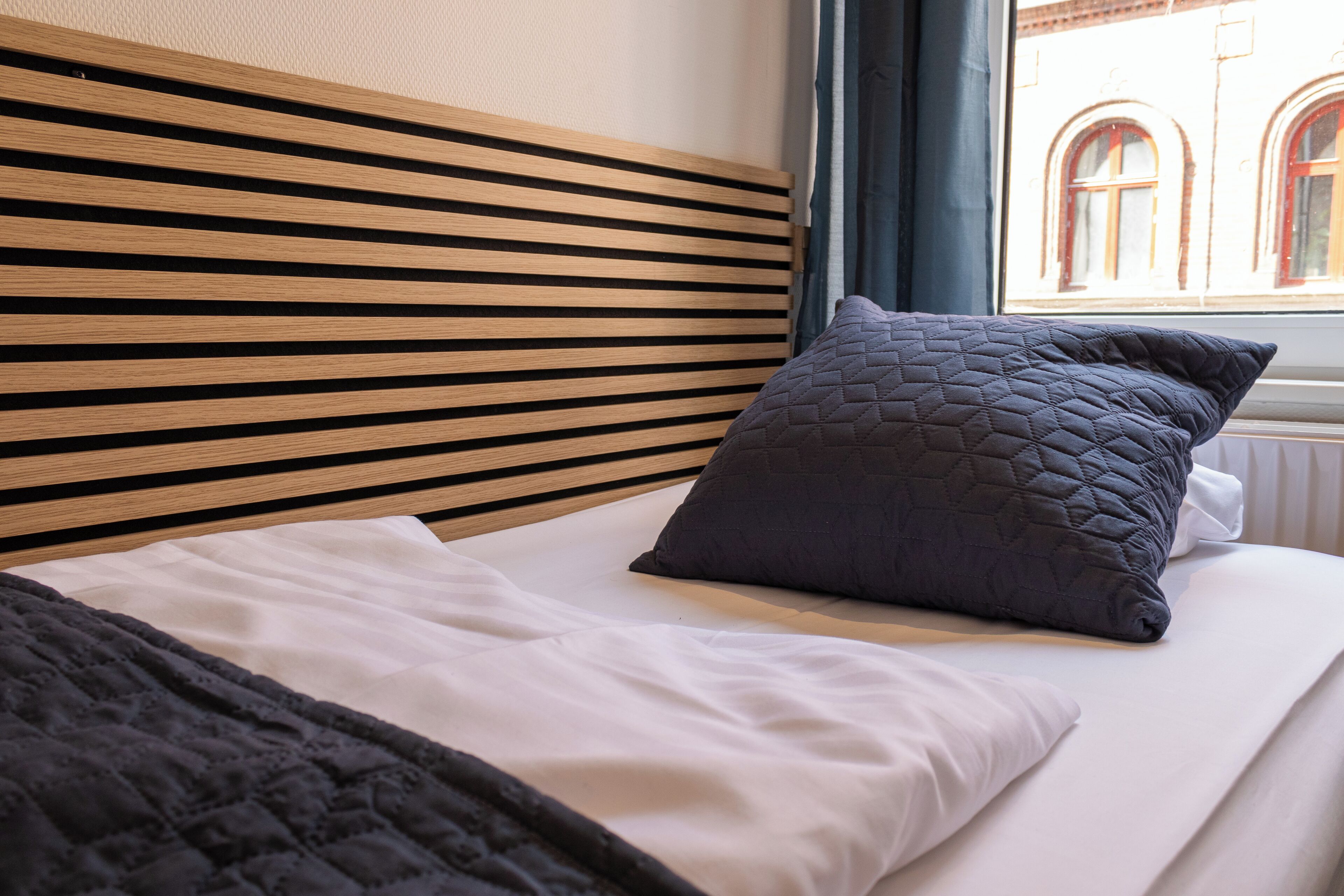 Een keurig opgemaakt bed met een gewatteerd kussen en een donkere deken, een houten hoofdbord en een raam met gordijnen.