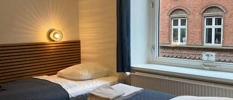 Habitación de hotel con dos camas, cabecero de madera, lámpara de pared y ventana con vista a un edificio de ladrillo.