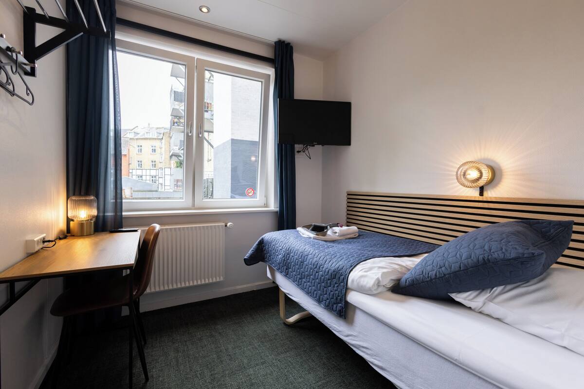 Habitación de hotel con cama, escritorio, silla, televisor montado en la pared y ventana con cortinas.