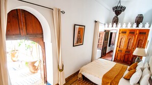 A bedroom with a large bed, a wooden wardrobe, a framed picture on the wall, and a doorway leading to an outdoor area with potted plants.