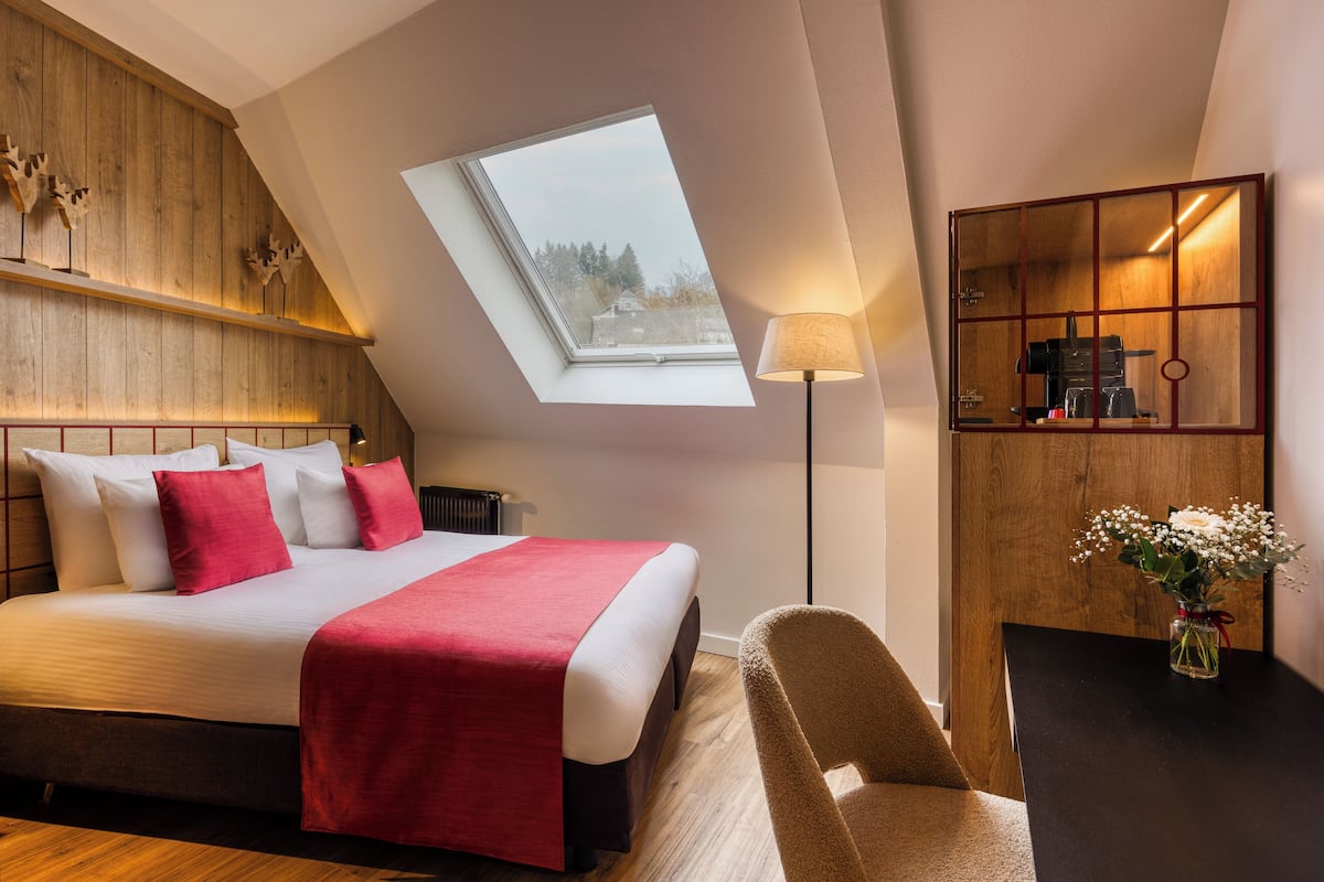 Un dormitorio con tragaluz, una cama con ropa de cama roja y blanca, un cabecero de madera, un escritorio con lámpara y un florero con flores.