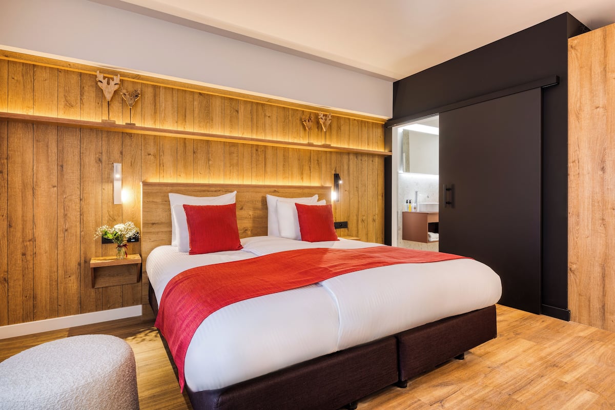 Un dormitorio moderno con cabecera de madera, una cama grande con ropa de cama roja y blanca, una mesita de noche con un florero y un armario negro.