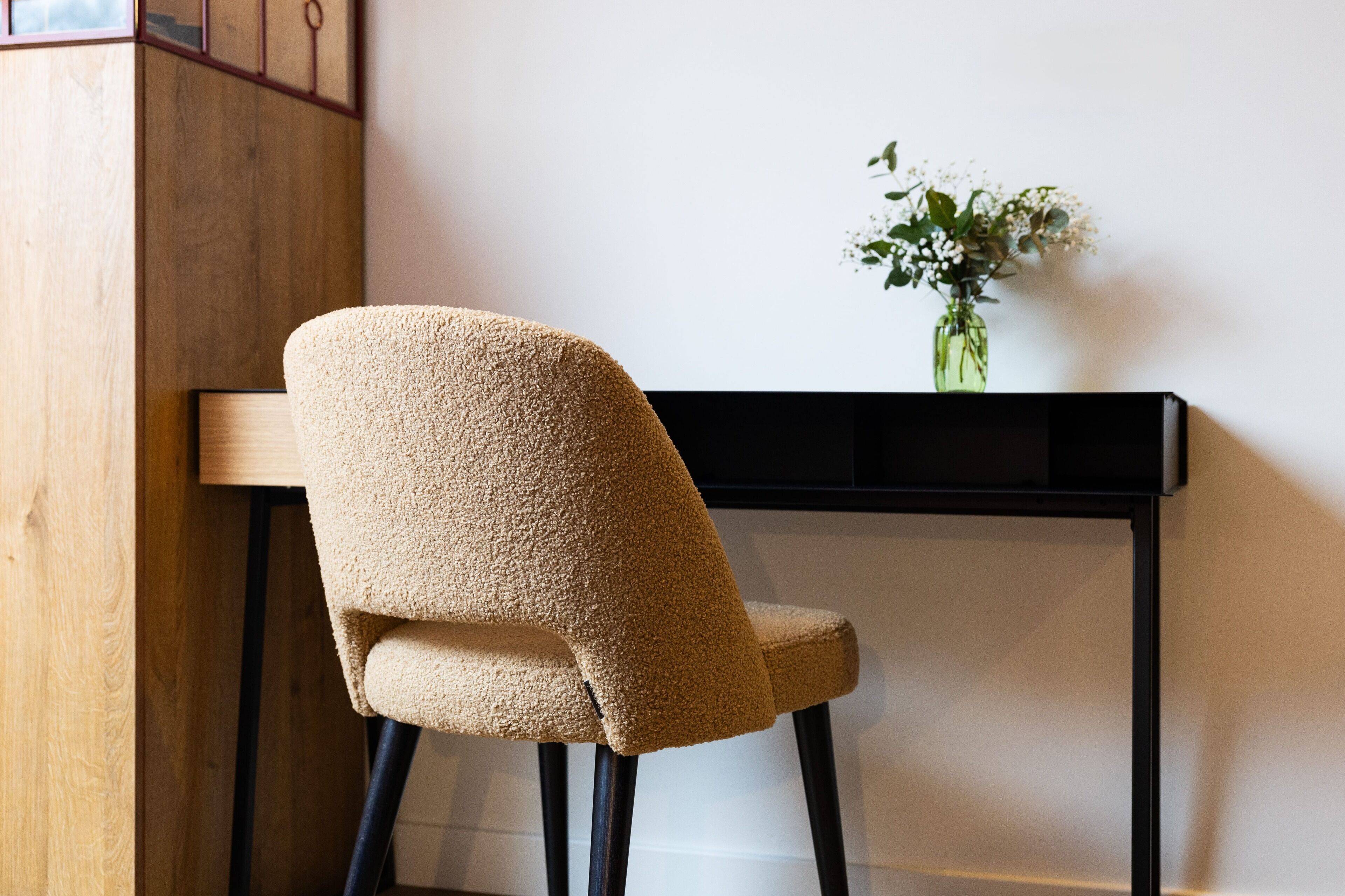 A chair with a textured fabric, a black table, and a vase with flowers on a white wall.