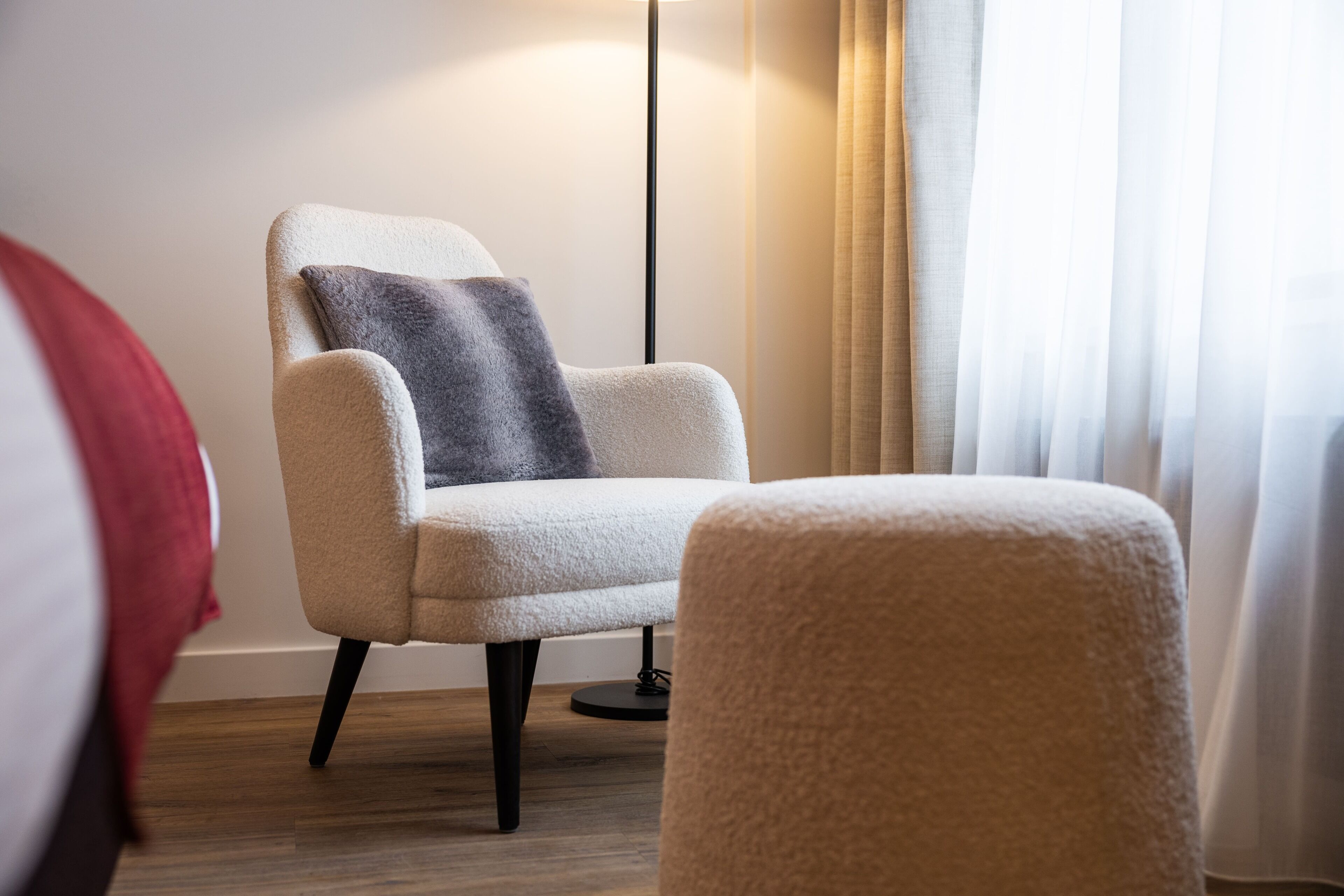 Habitació d'hotel moderna amb un butaca confortable, una làmpada de peu i una finestra gran amb cortines translúcides.