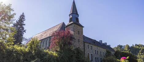 A historic stone building with a prominent tower and a weather vane, surrounded by lush greenery and flowering plants.