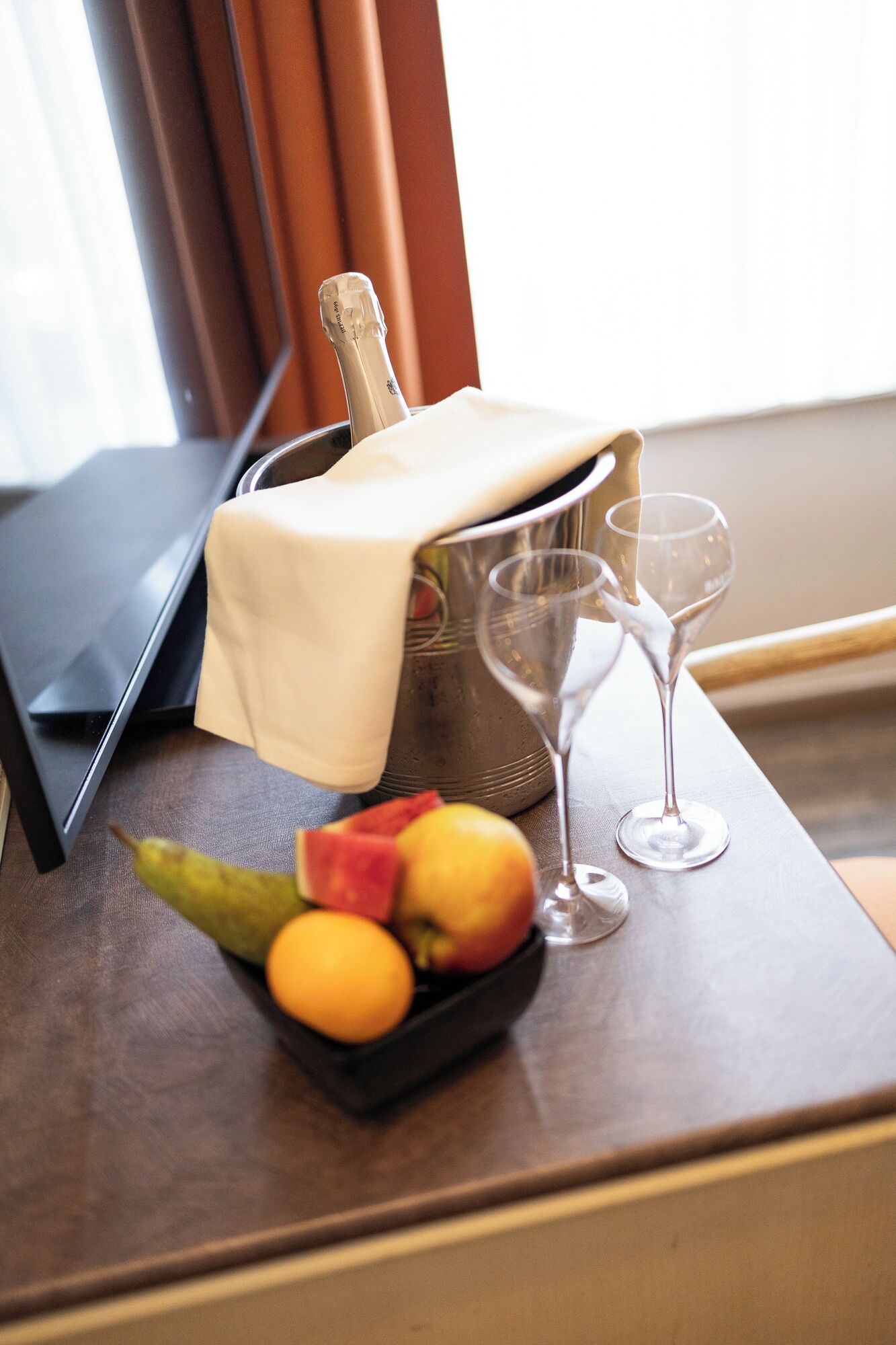 A hotel room with a laptop, a bottle of champagne, two wine glasses, and a bowl of fruit on the table.