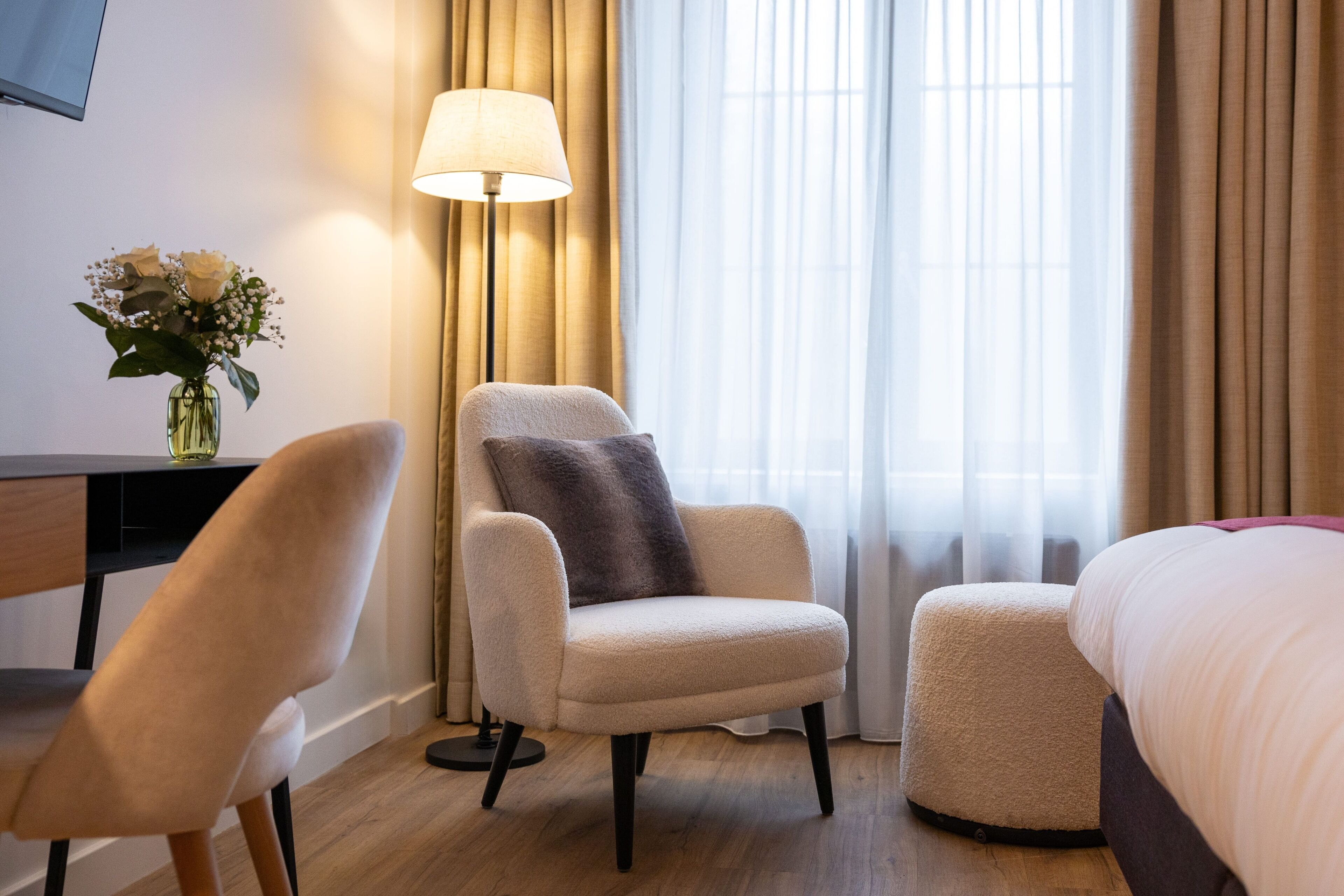 A hotel room with a bed, a desk, a chair, a lamp, and a vase with flowers.