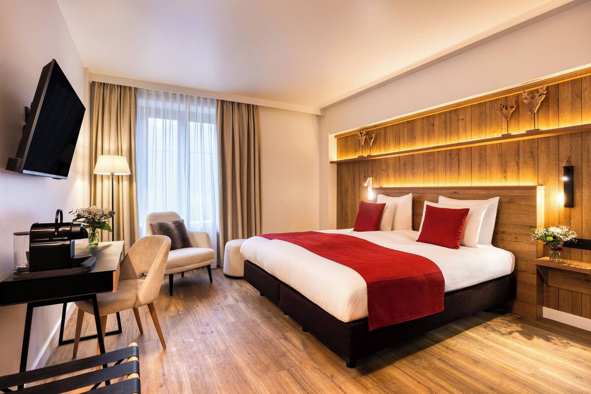 Habitación de hotel con una cama grande, un escritorio con silla, un televisor montado en la pared y un cabecero de madera.