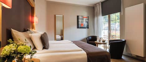A hotel room with a large bed, a desk with a chair, a small table with a chair, a large window with curtains, and a painting on the wall.