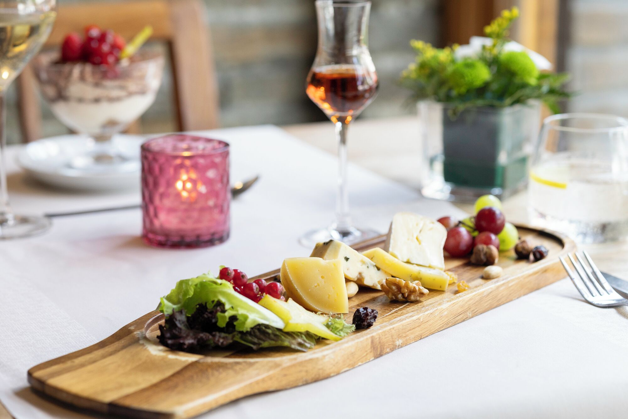En træplanke med ost, druer, nødder og en salat på et bord med et lys og glas.