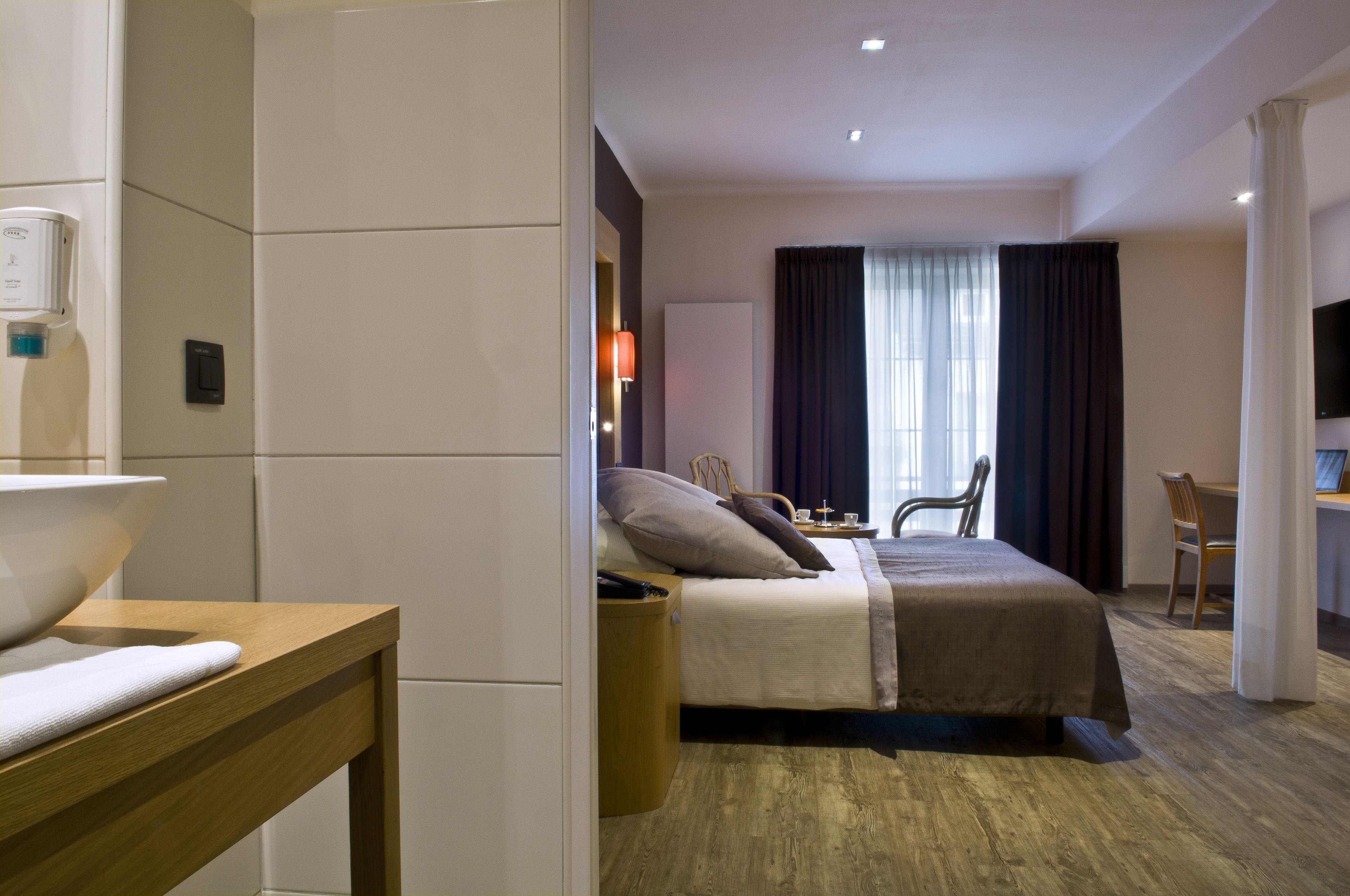 A hotel room with a bed, a desk, a chair, a TV, and a bathroom area with a sink and mirror.