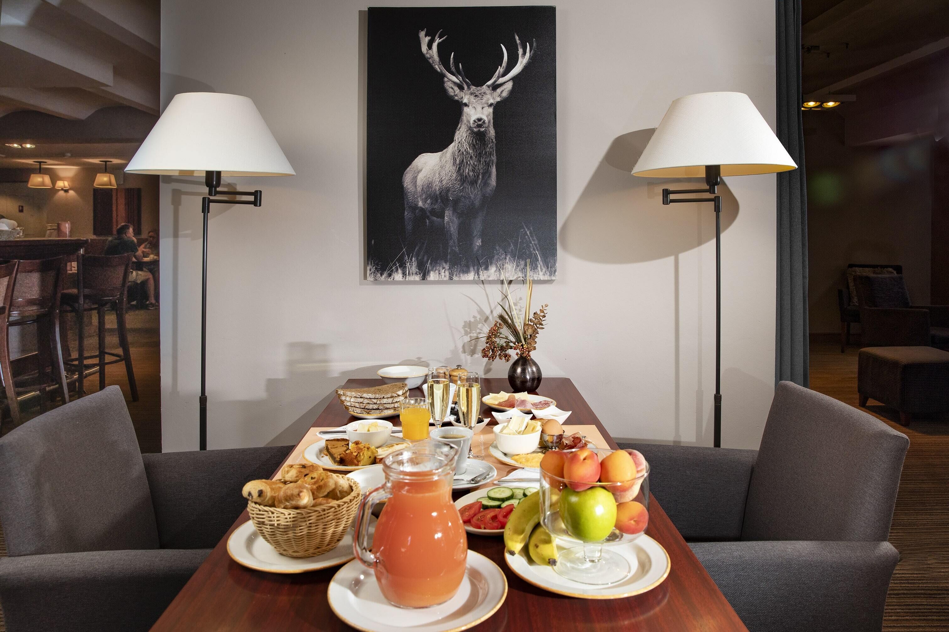 A dining table set with breakfast items, a painting of a deer, and two floor lamps.