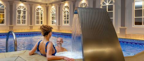 An indoor swimming pool with a unique water feature, a tiled floor, and arched windows.