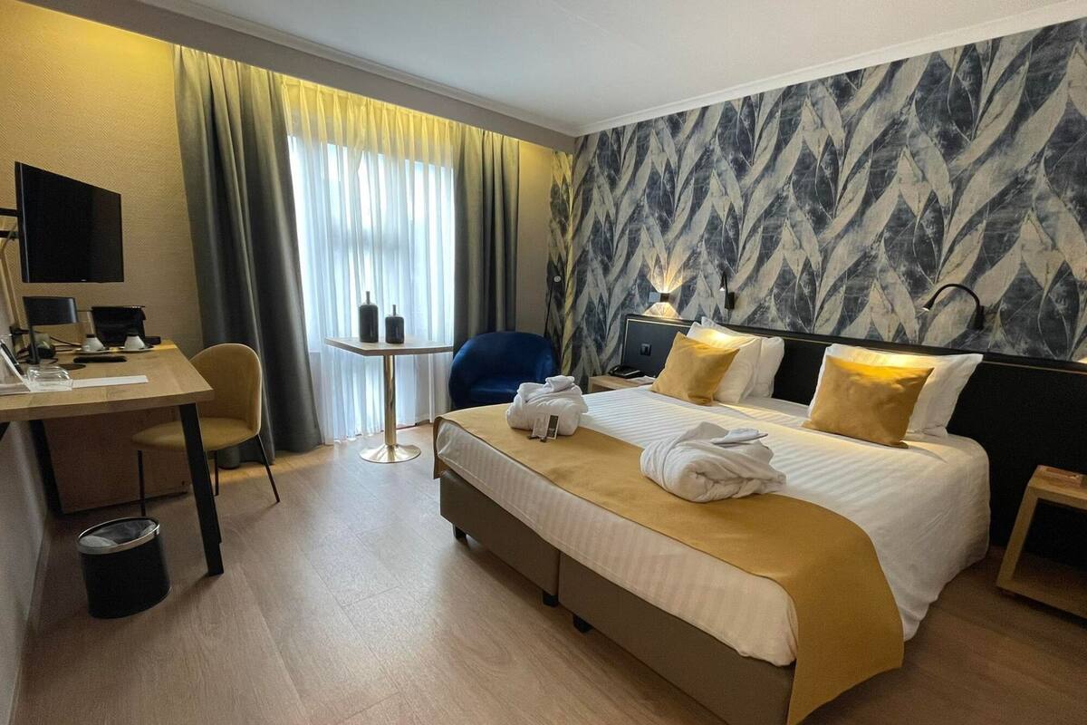 A hotel room with a bed, a desk, a chair, a small table, and a trash bin.