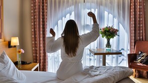 A person sitting on a bed in a hotel room with a window, a bedside table with a lamp, and a chair. - Hotel Quartier Latin (Marche-en-Famenne)