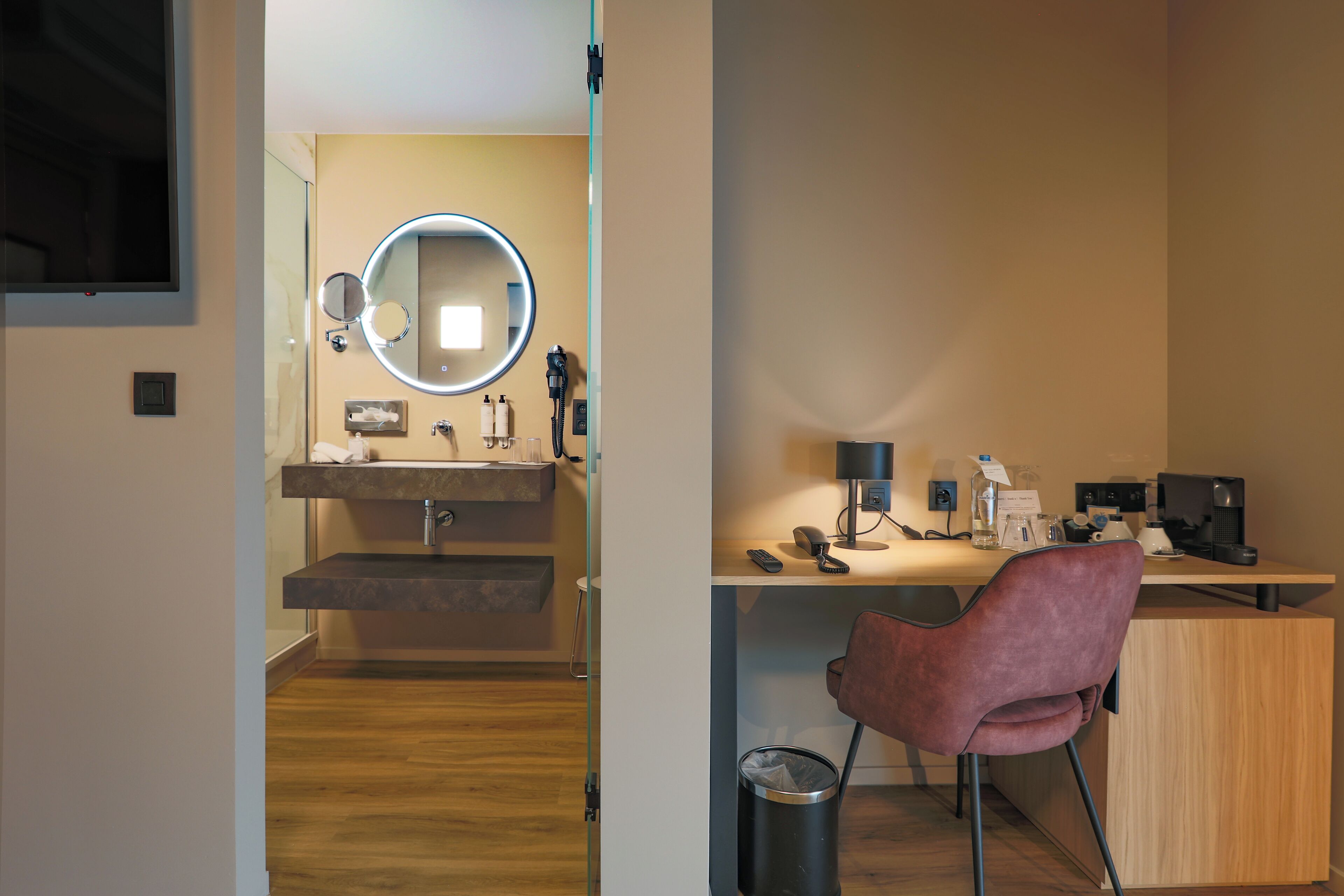 A modern bathroom with a mirror, sink, and vanity. There is a desk with a chair and a trash can.