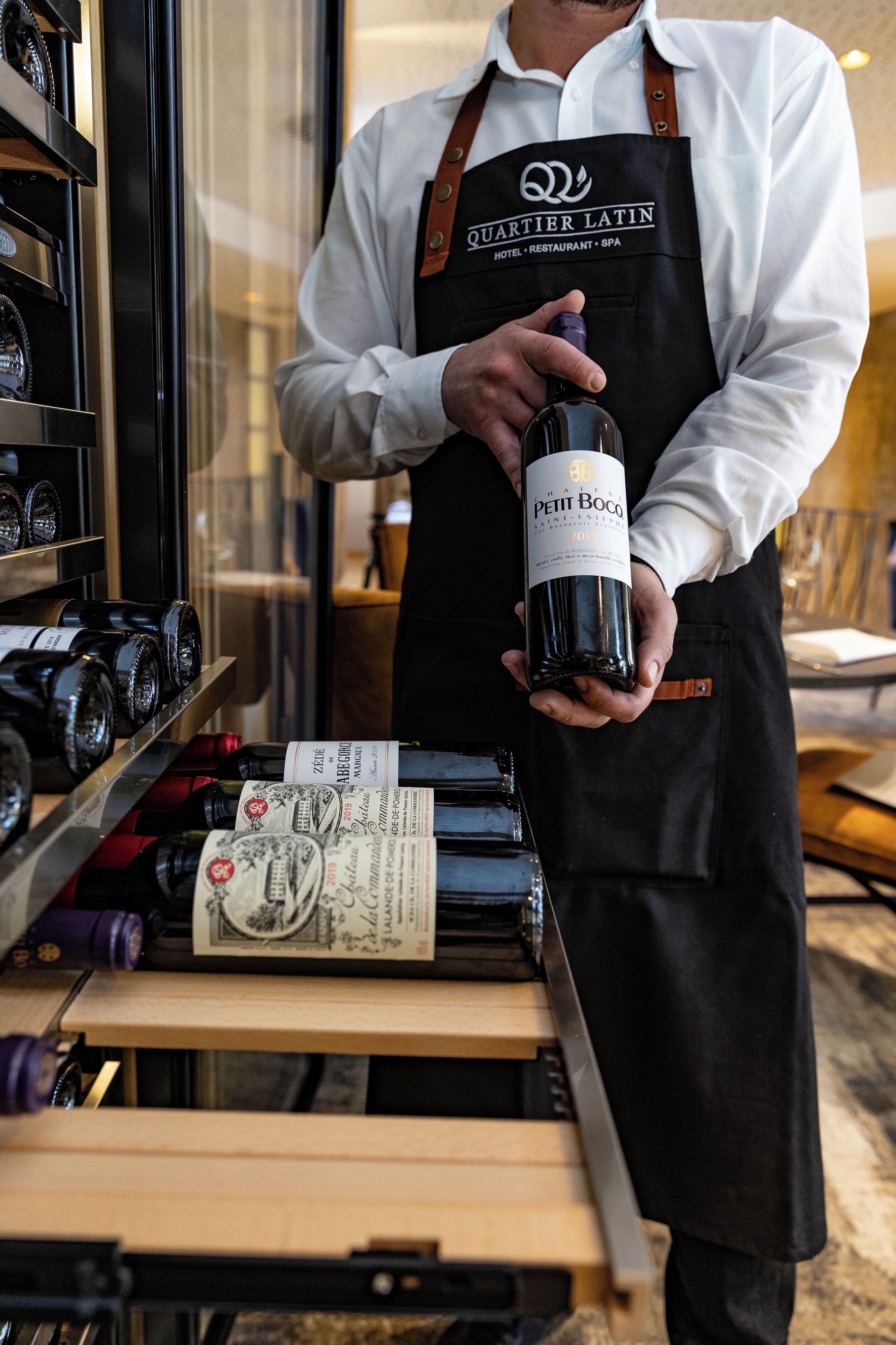 A person in a white shirt and black apron holding a bottle of wine at a counter with wine bottles and price tags.