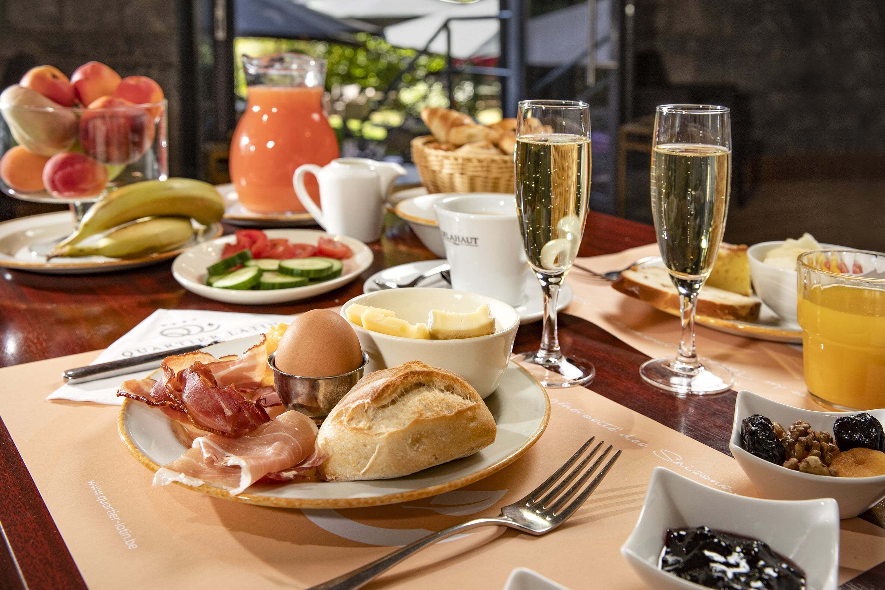Une table de petit-déjeuner avec des croissants, du bacon, des œufs et des fruits. On y trouve également deux verres de champagne et une carafe de jus d’orange.