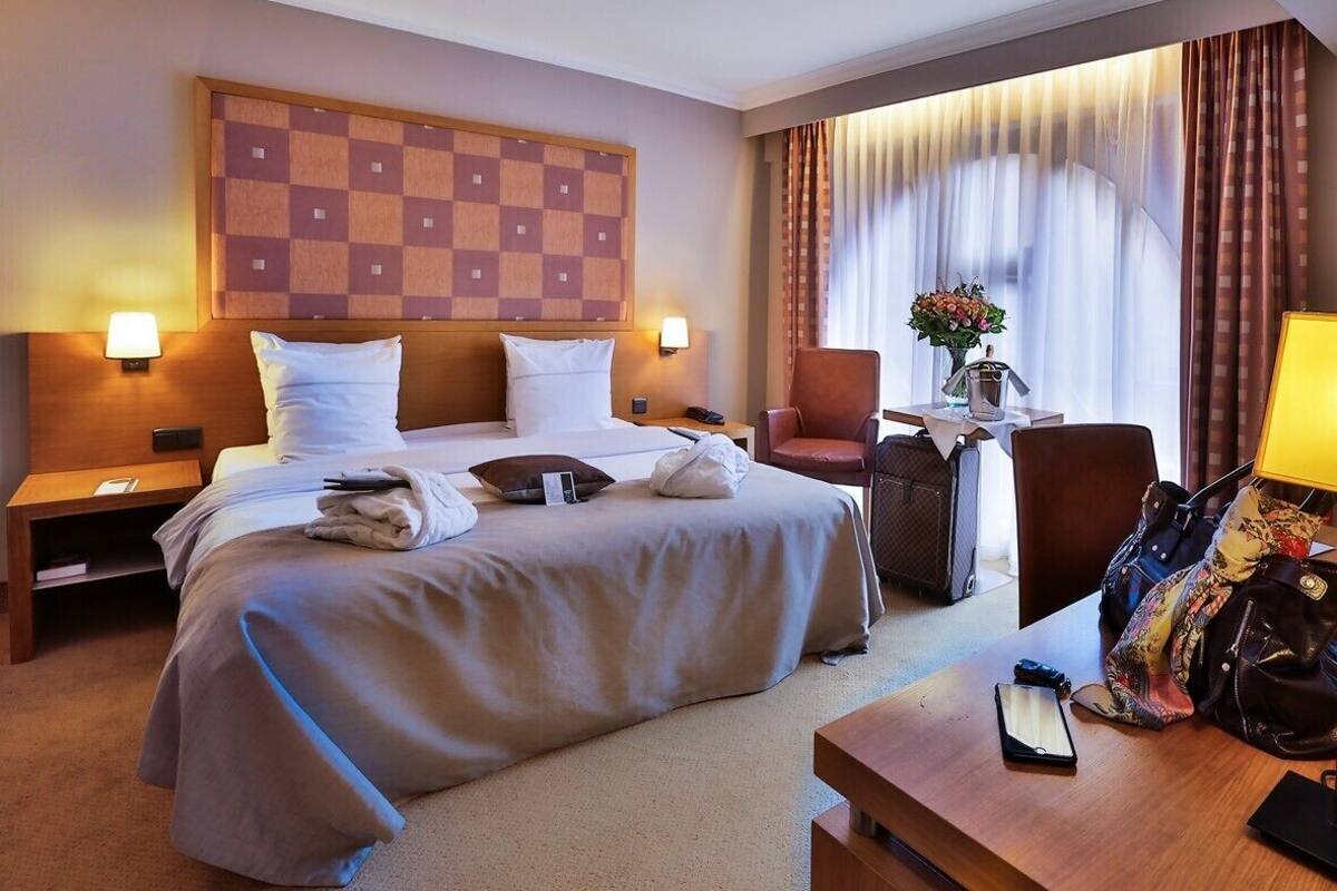 A hotel room with a large bed, two bedside tables with lamps, a desk with a chair, and a window with curtains.