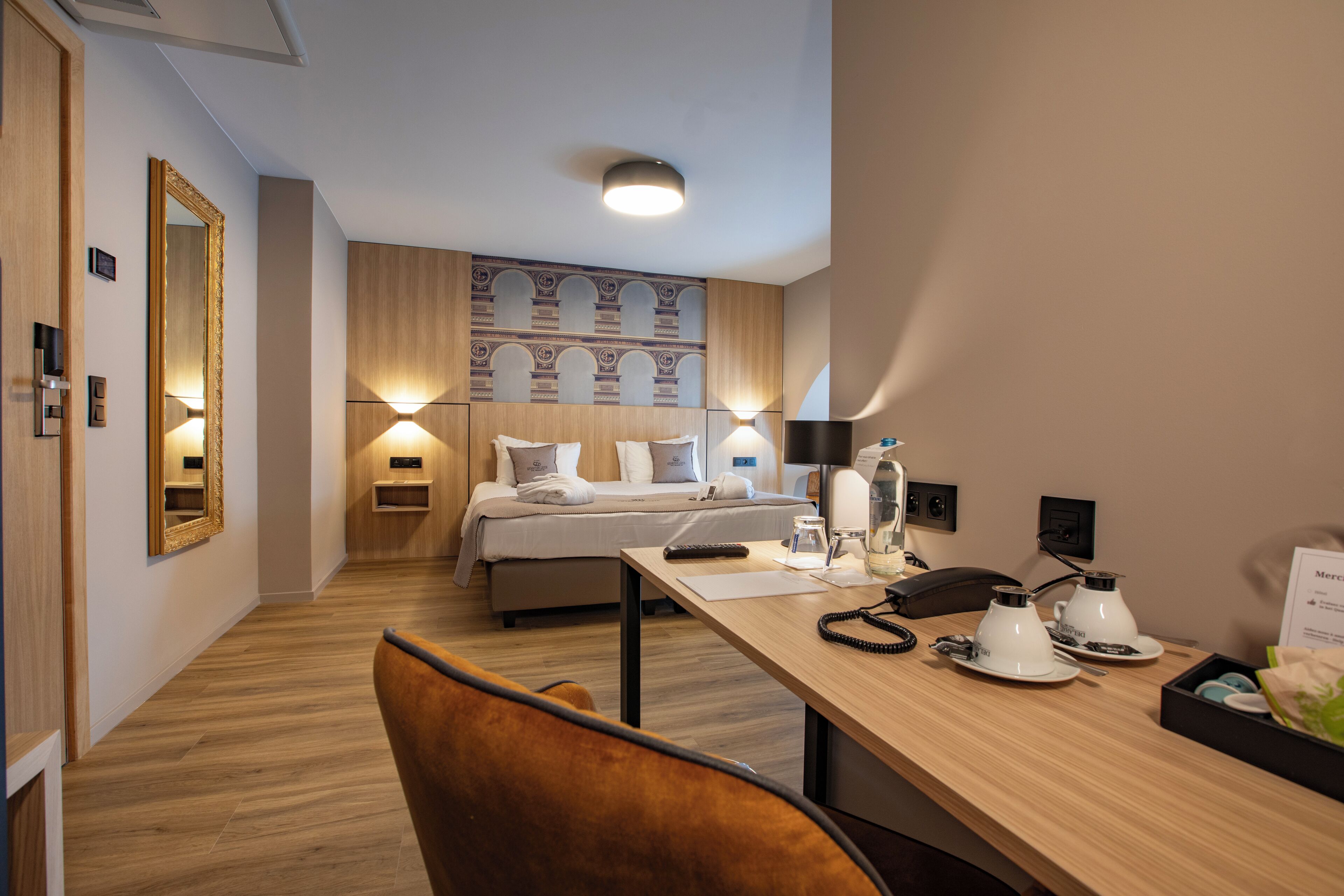 A hotel room with a bed, bedside tables, a desk with a chair, a mirror, and a wall-mounted light fixture.