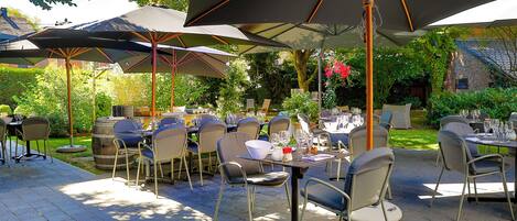 Patio esterno con tavoli e sedie sotto grandi ombrelloni, circondato da vegetazione e un edificio in mattoni.